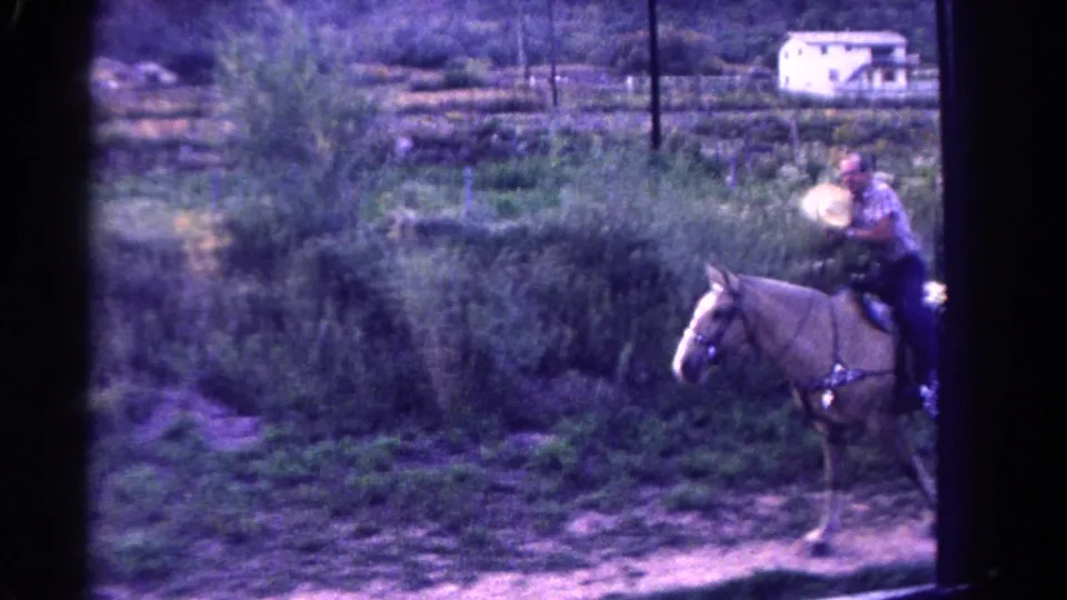 1967 Colorado home-movie frame showing a man on a light-colored horse moving past brush, with a small white structure in the background.