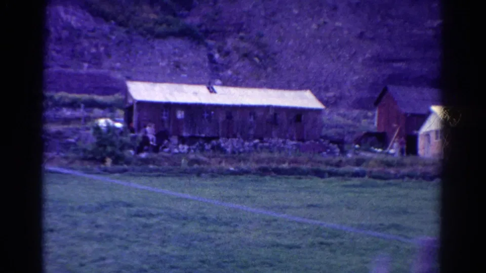 1967 Colorado frame with the long wooden building, nearby tiny figures, a small tree, pasture, and part of another outbuilding at right.