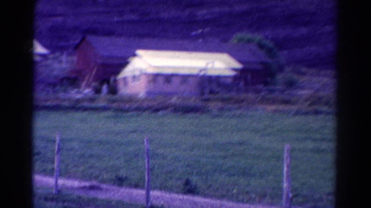 1967 Colorado home-movie frame showing a distant rural house, green field, fence posts, and dark mountains beyond.