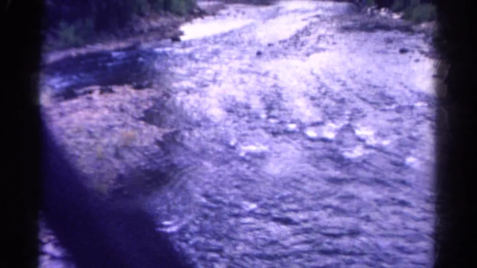Shallow rocky Colorado stream filmed from a moving bridge or vehicle, with water reflecting light across the stones.