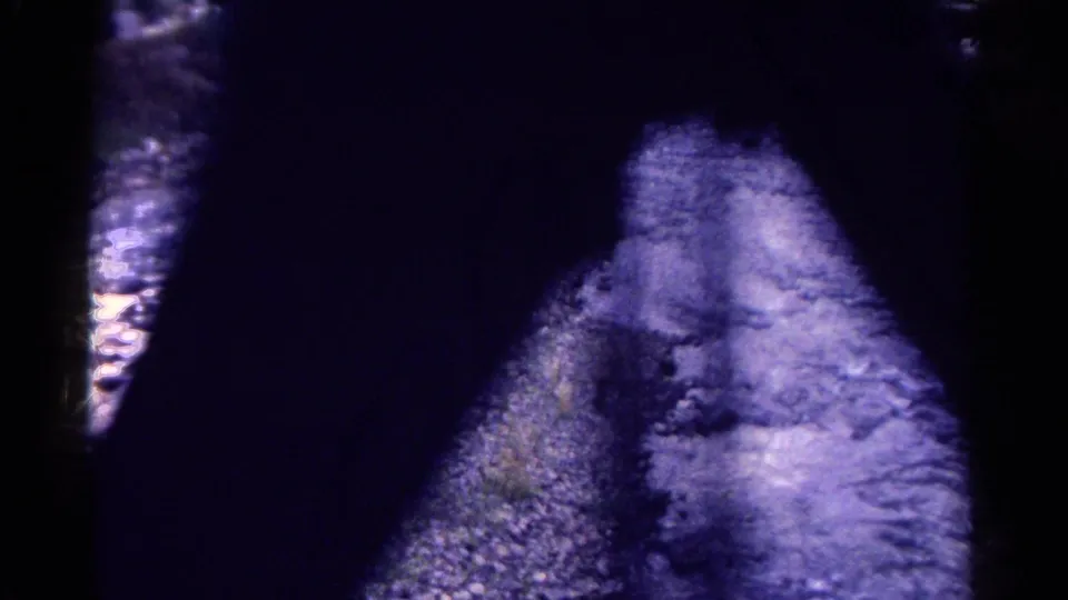 A dark, heavily shadowed bridge passage reveals a narrow bright section of rocky water and stream bank below.