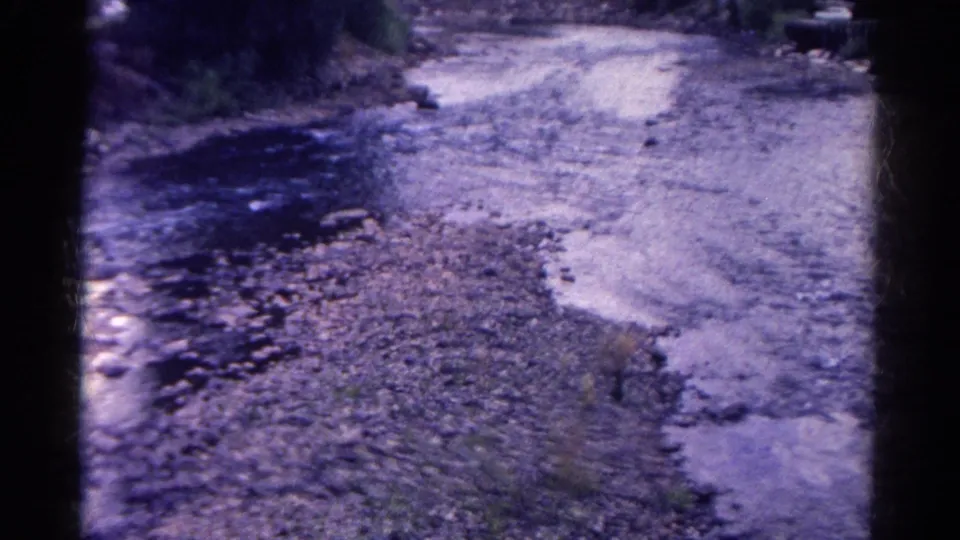 1967 Colorado 8mm frame showing a rocky streambed with shallow flowing water and a rough roadside or bank above it.