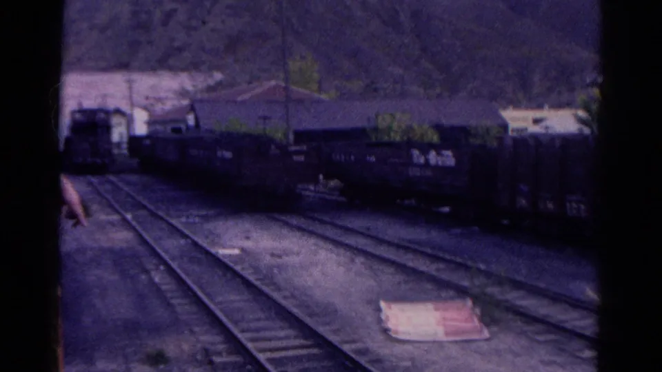 1967 Colorado 8mm view of a rail yard with curving tracks, freight cars, low buildings, hills, and a partly visible person at frame edge.