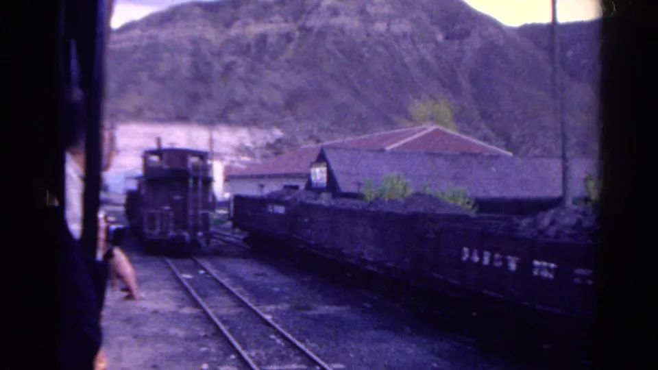 1967 Colorado rail-yard frame showing a small locomotive, dark freight cars, converging tracks, a low building, utility pole, and hillside beyond.