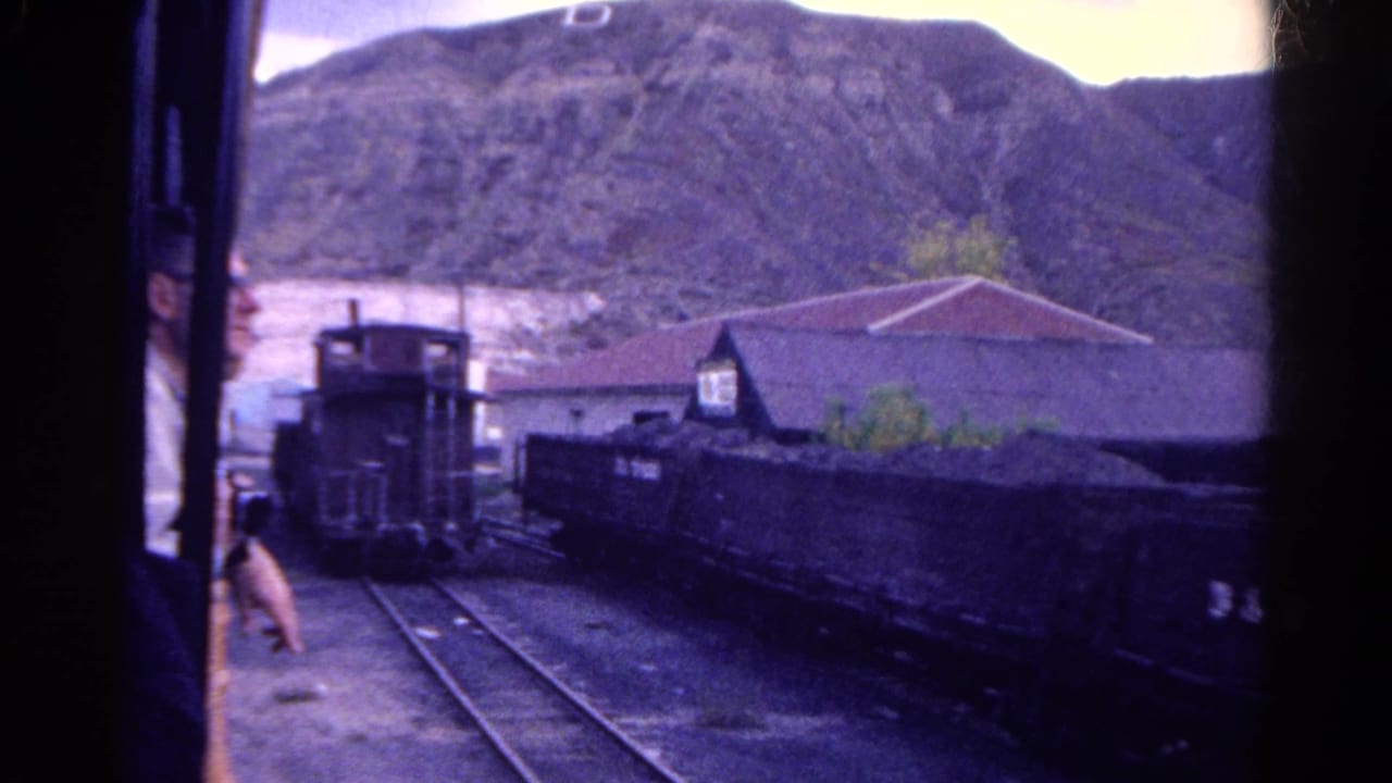 Passenger train window view of a rail yard with freight cars, caboose, rails, building, and rocky hill.