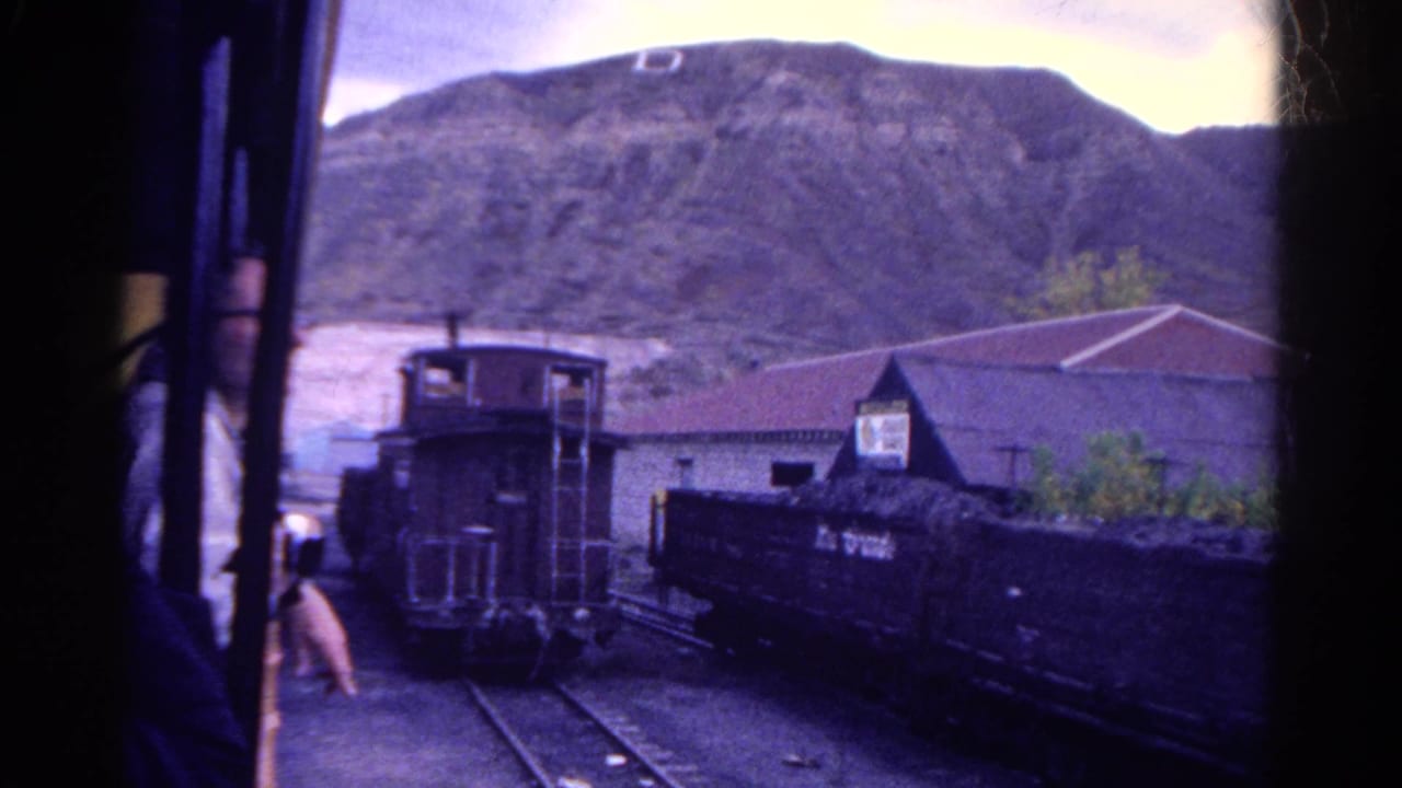 A passenger in a 1967 Colorado train car points out the window toward a rail yard with freight cars, a caboose, and rocky hillside beyond.