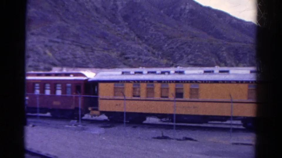 A 1967 Colorado 8mm view of yellow and red passenger rail cars beside a fenced rail area and rocky mountain backdrop.