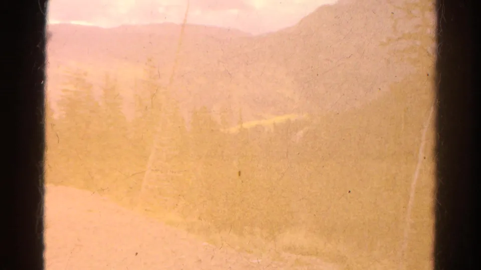 Faded 1967 Colorado mountain and valley view with bare trees, warm haze, dark frame edge, and visible 8mm surface wear.