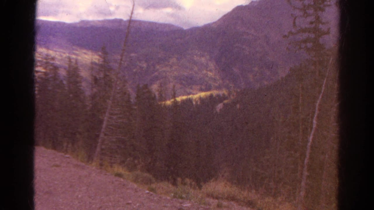1967 Colorado 8mm frame showing pine-covered slopes, distant mountains, and a dirt foreground edge with dark film-gate borders.