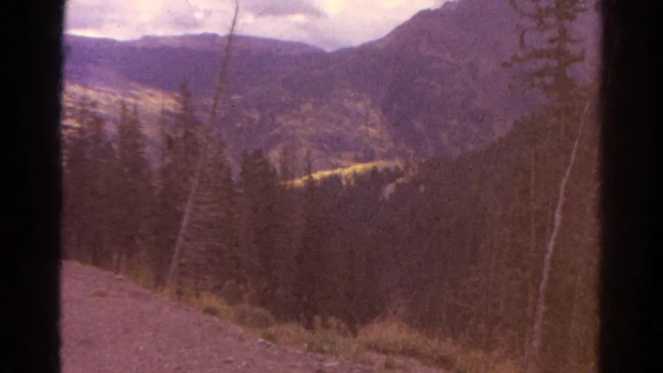 1967 Colorado 8mm frame showing pine-covered slopes, distant mountains, and a dirt foreground edge with dark film-gate borders.