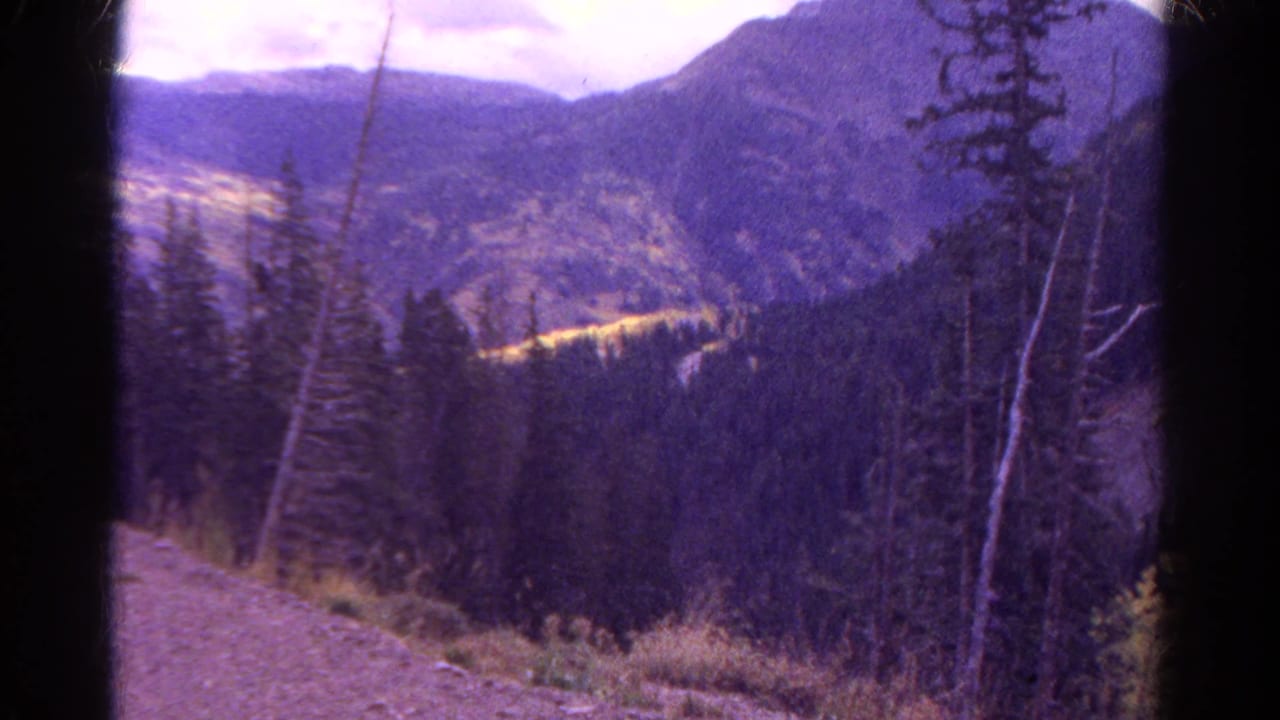 1967 Colorado home-movie frame with purple color shift, pine-covered valley, distant ridges, and dark gate edges.