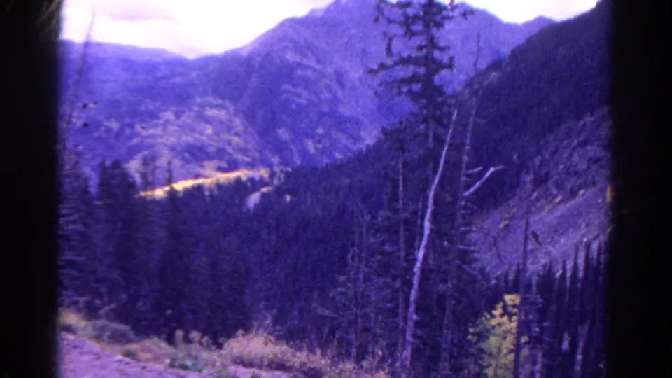 1967 Colorado home-movie frame with evergreen trees, rugged slopes, a distant valley, and purple color cast from aged film.