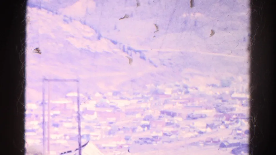 1967 Colorado 8mm frame showing a distant town in a valley beneath pale mountain slopes, with utility poles near the foreground.