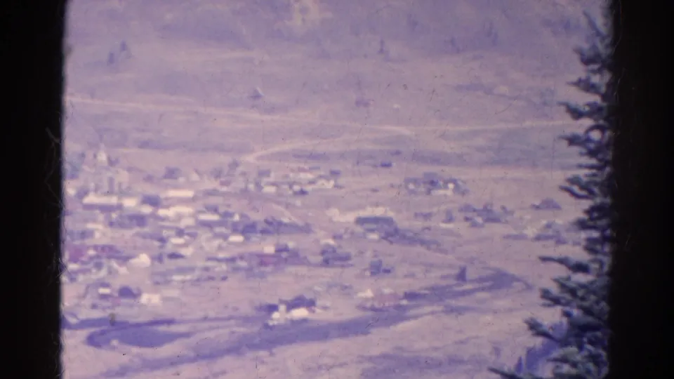 1967 Colorado mountain-town view with a winding road, scattered buildings, open hillside, and a pine at the right edge.