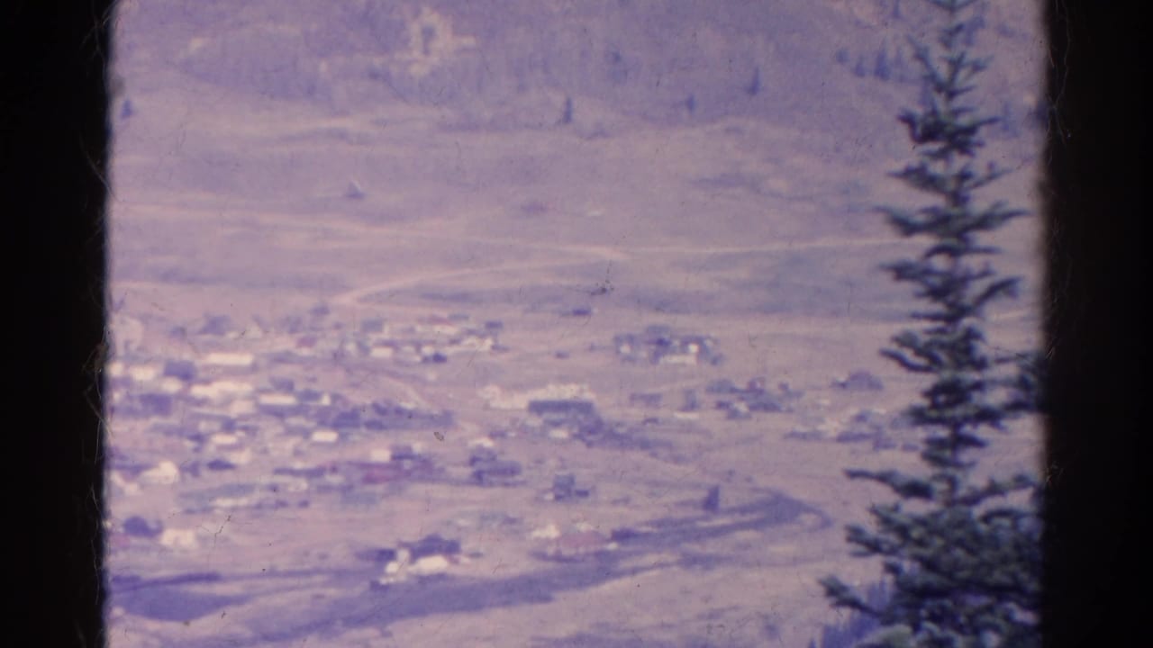 Distant settlement and dry hillside seen past a dark vertical film edge and evergreen tree.
