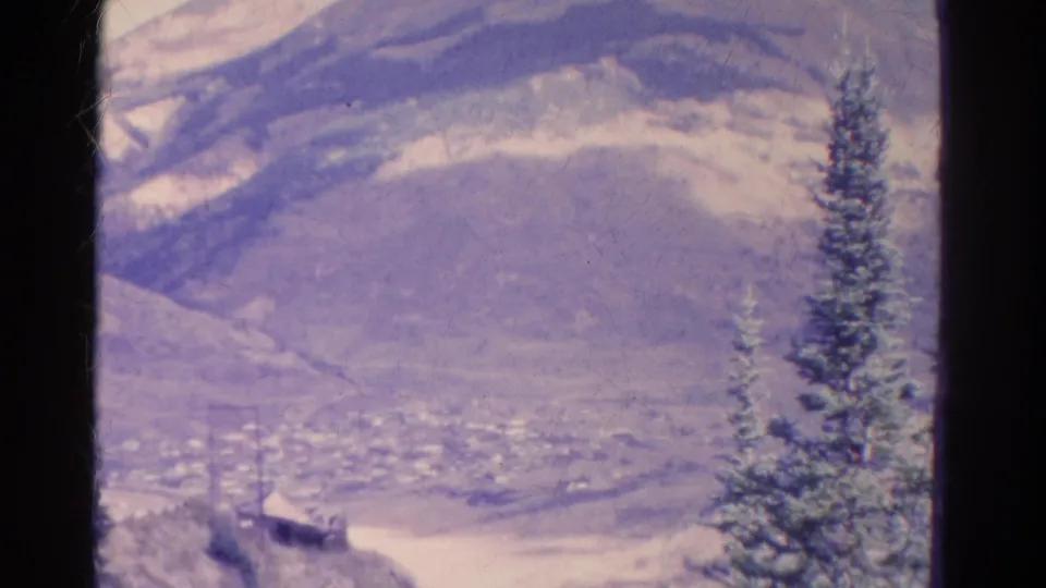 1967 Colorado frame showing a distant town beneath a broad dry hillside, with an evergreen tree framing the right side.