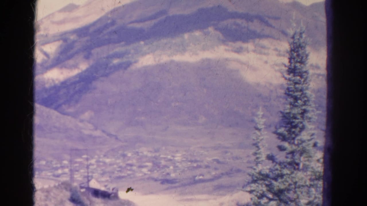 1967 Colorado home-movie frame showing a hazy mountain valley, distant town, and foreground evergreen within dark film borders.