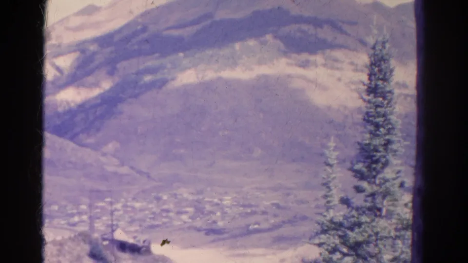 1967 Colorado home-movie frame showing a hazy mountain valley, distant town, and foreground evergreen within dark film borders.