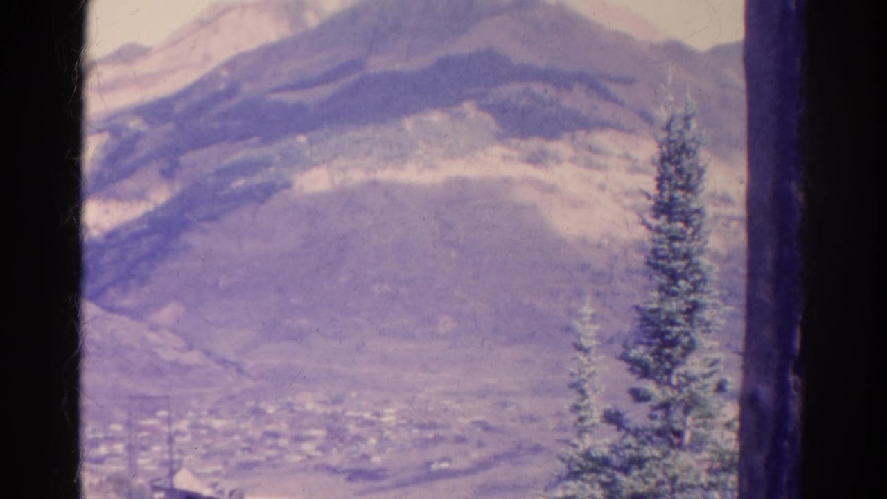 1967 Colorado scenic frame with layered mountain ridges, a faint valley settlement, and a prominent evergreen in the foreground.