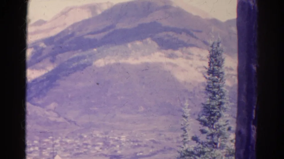 Soft 1967 Colorado home-movie frame of a mountain valley, faint town structures, and an evergreen beside dark film borders.
