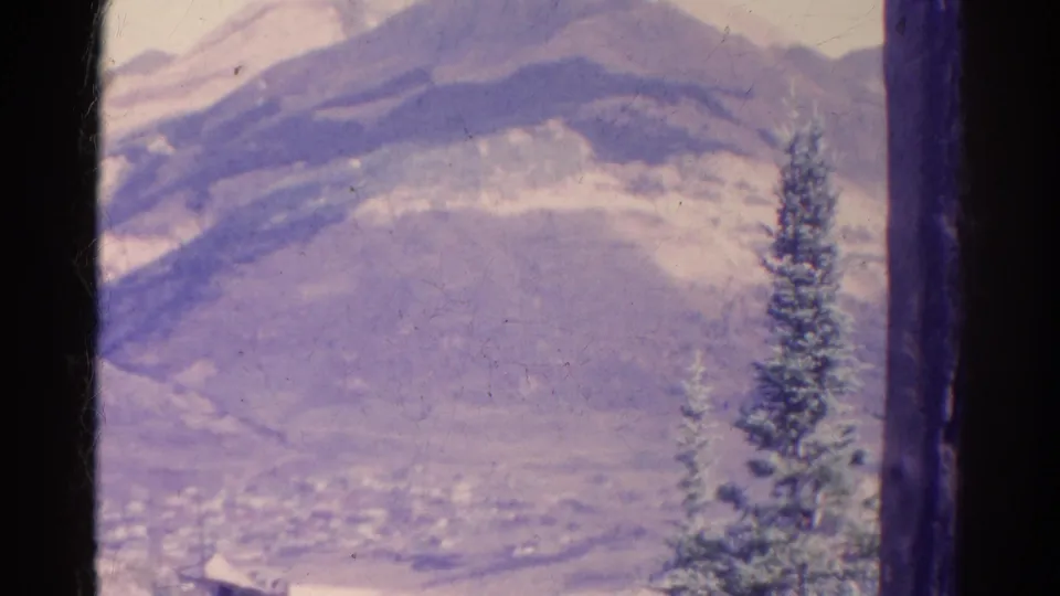 1967 Colorado 8mm frame showing a hazy mountain valley, evergreen foreground, and small distant town shapes under a bright sky.