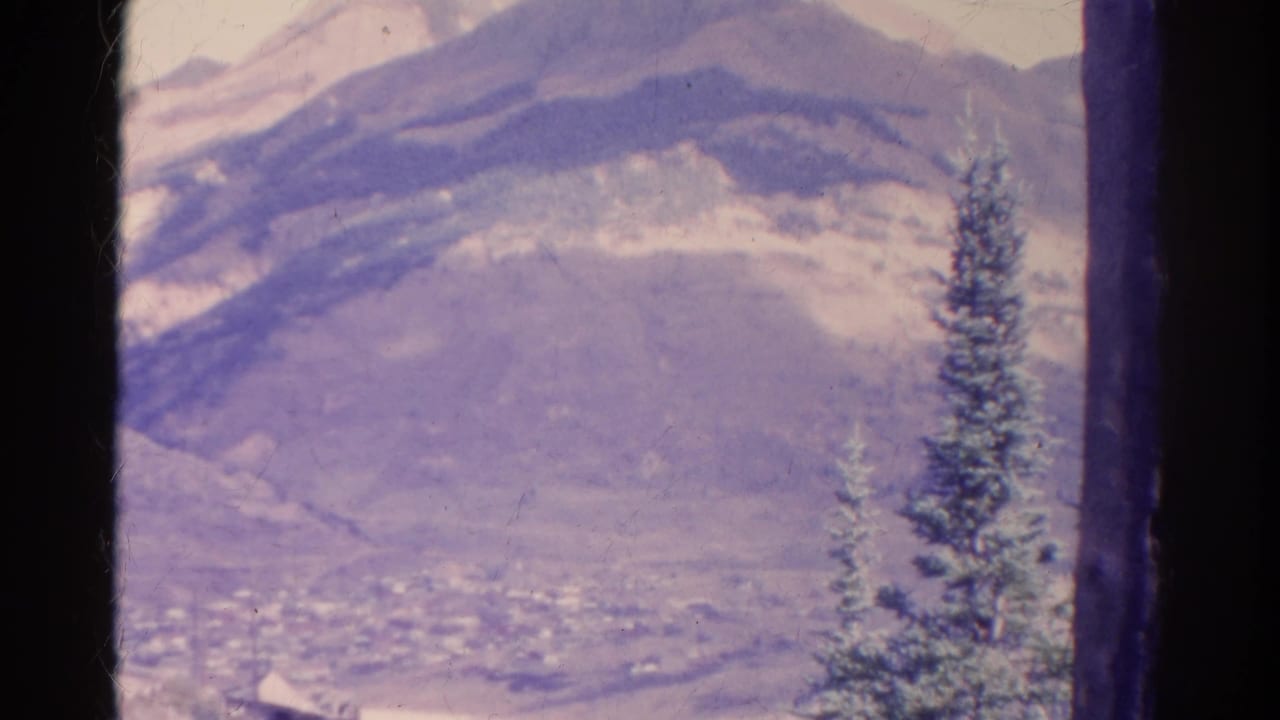1967 Colorado 8mm frame showing a snow-dusted mountain landscape, distant valley, dark gate edges, and a tall evergreen in the foreground.