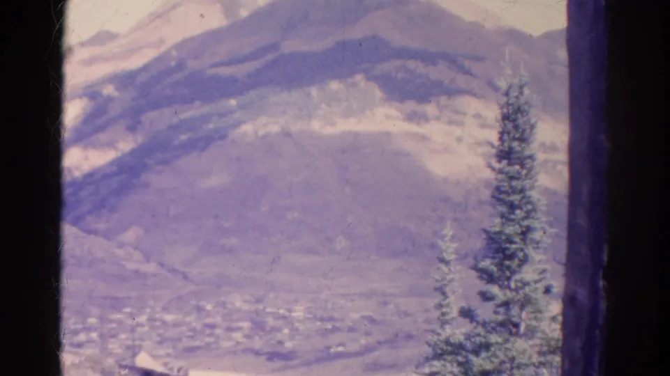 1967 Colorado 8mm frame showing a snow-dusted mountain landscape, distant valley, dark gate edges, and a tall evergreen in the foreground.