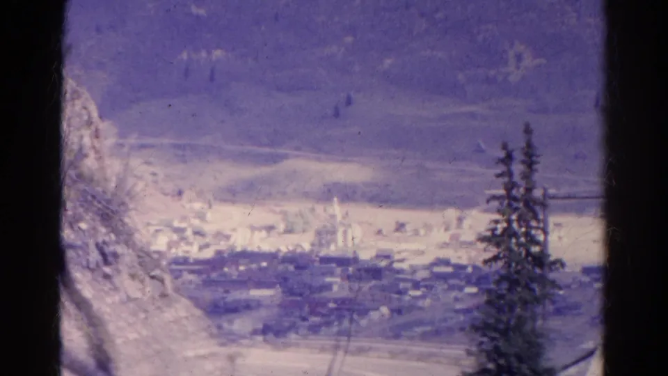 1967 Colorado 8mm frame showing a distant snowy valley town below mountain slopes, framed by evergreens and dark film edges.
