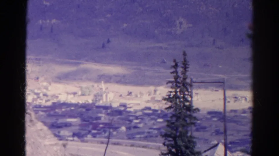 1967 Colorado home-movie frame looking over a small town between pale mountain slopes, framed by trees, pole lines and film edges.