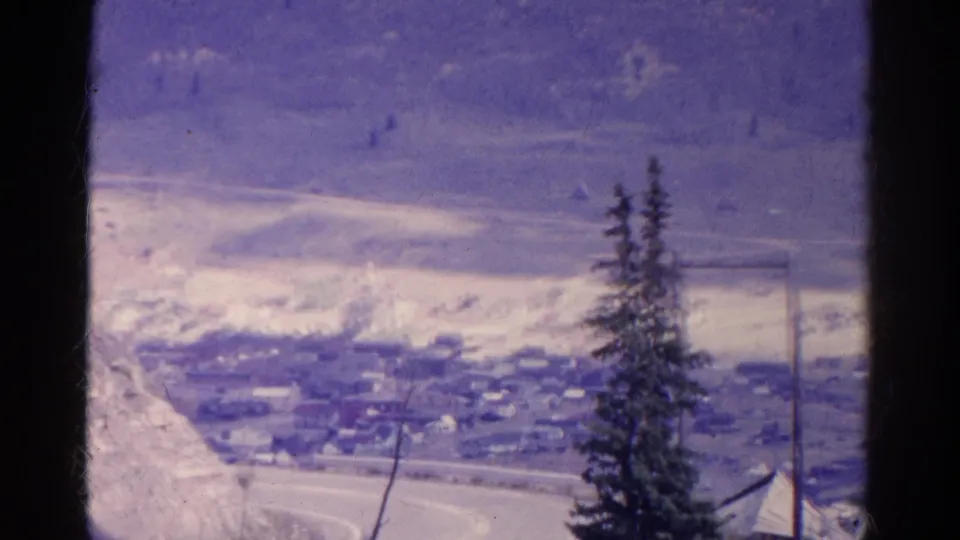 1967 Colorado 8mm still showing a winding road and clustered buildings below an overlook, with an evergreen tree in the foreground.