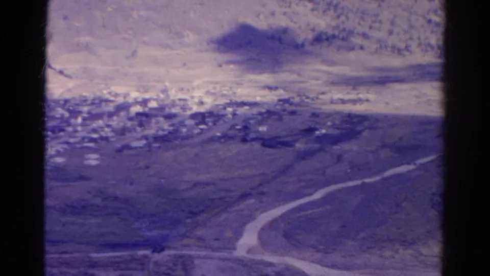 1967 Colorado 8mm frame with a pale winding road, scattered town structures and mountain slopes under a purple-tinted sky.