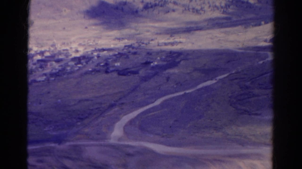 Aerial view over barren land with a winding road running toward a distant cluster of buildings.