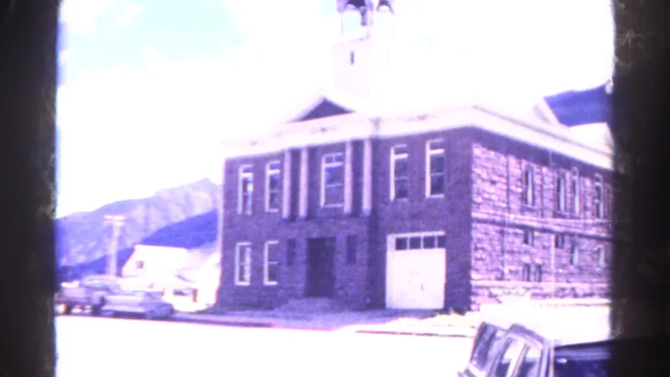 Overexposed 1967 Colorado street view of a stone building with bell tower, mountain backdrop, and parked car.