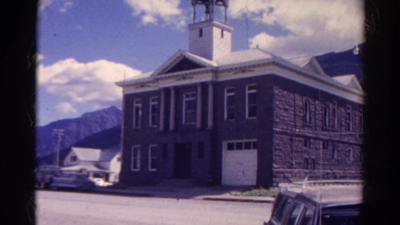 1967 Colorado 8mm frame showing a large stone civic-looking building with columns, bell tower, parked cars, mountains, and blue sky.