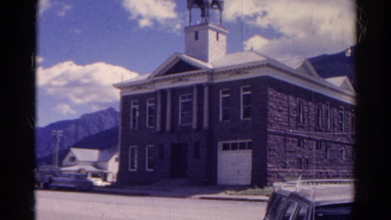 Stone building with bell tower, columns, parked cars, and mountain backdrop.