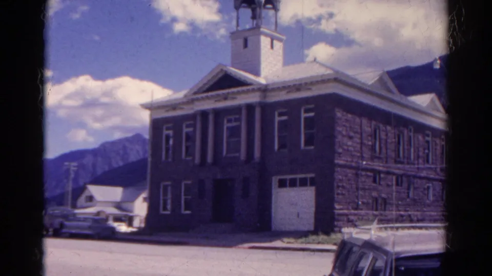Soft 8mm frame from Colorado in 1967 showing a grand stone building, tower, cars, curb, and rugged mountain background.