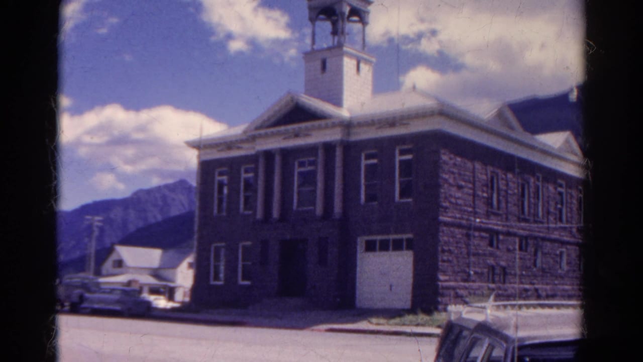 Similar view of a stone bell-tower building with cars, mountains, and bright sky.
