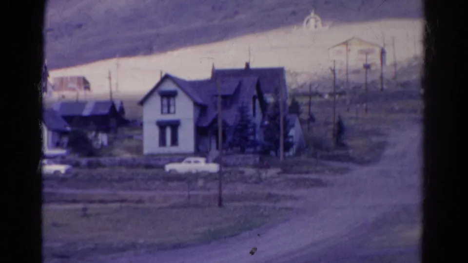 1967 Colorado 8mm view of roadside houses, a parked white car, utility poles, open ground, and a dirt road curving past the homes.