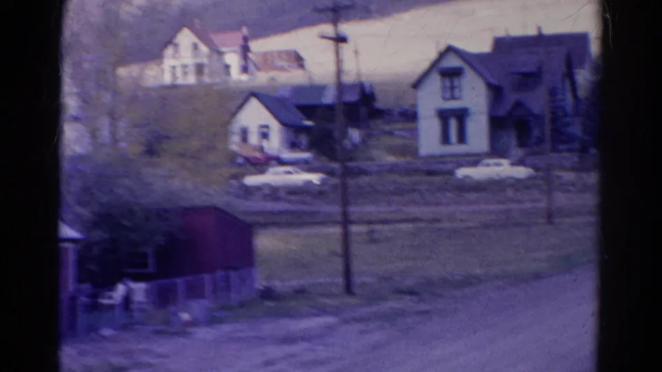 1967 Colorado 8mm frame showing several rural house exteriors, parked cars, utility poles, a foreground road, and a pale hillside behind them.