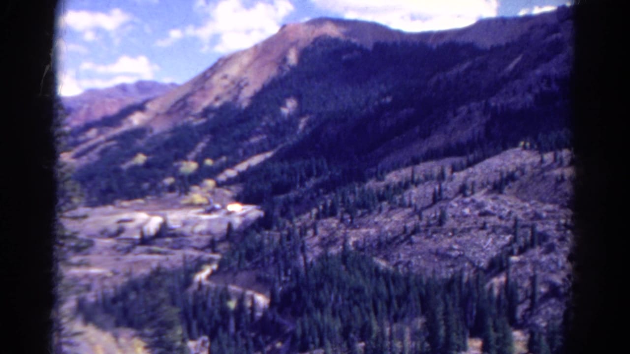 1967 Colorado home-movie frame with evergreen-covered slopes, pale valley terrain, and a high mountain ridge in soft 8mm color.
