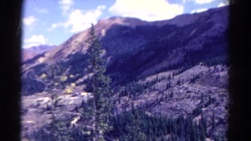 1967 Colorado home-movie frame showing a foreground evergreen against a broad mountain valley and blue-purple tinted ridgeline.