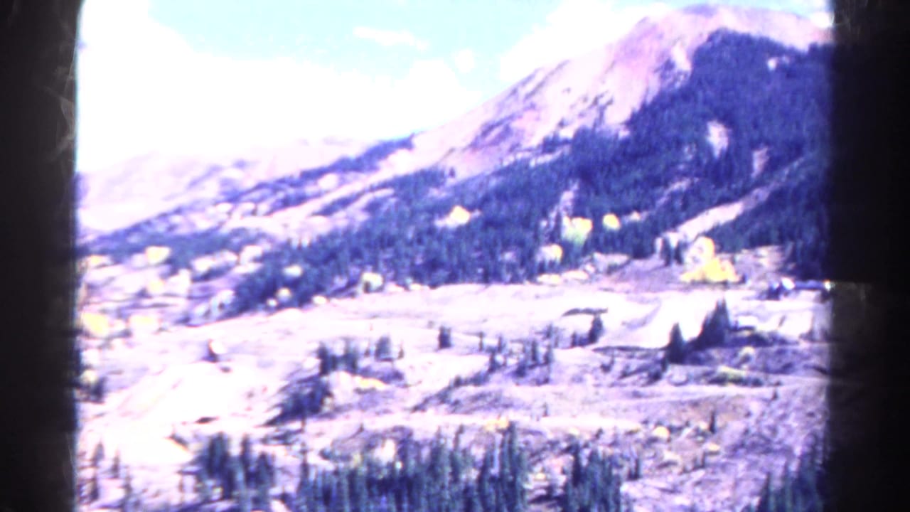 Wide mountain valley with forested slopes, bright sky, and cleared work-site ground below.