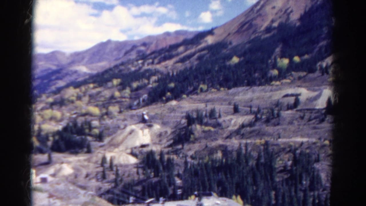Colorado mountain landscape on 8mm film, with unpaved roads, forest patches, graded terrain, and small mining-site structures.