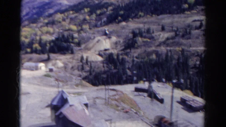 1967 Colorado home-movie frame showing a small roofed building, utility poles, roads, trees, and mountain work-site terrain.