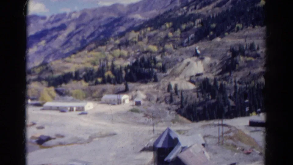 Soft 8mm Colorado valley frame with a small building in front, winding roads, scattered trees, and distant mountain slopes.