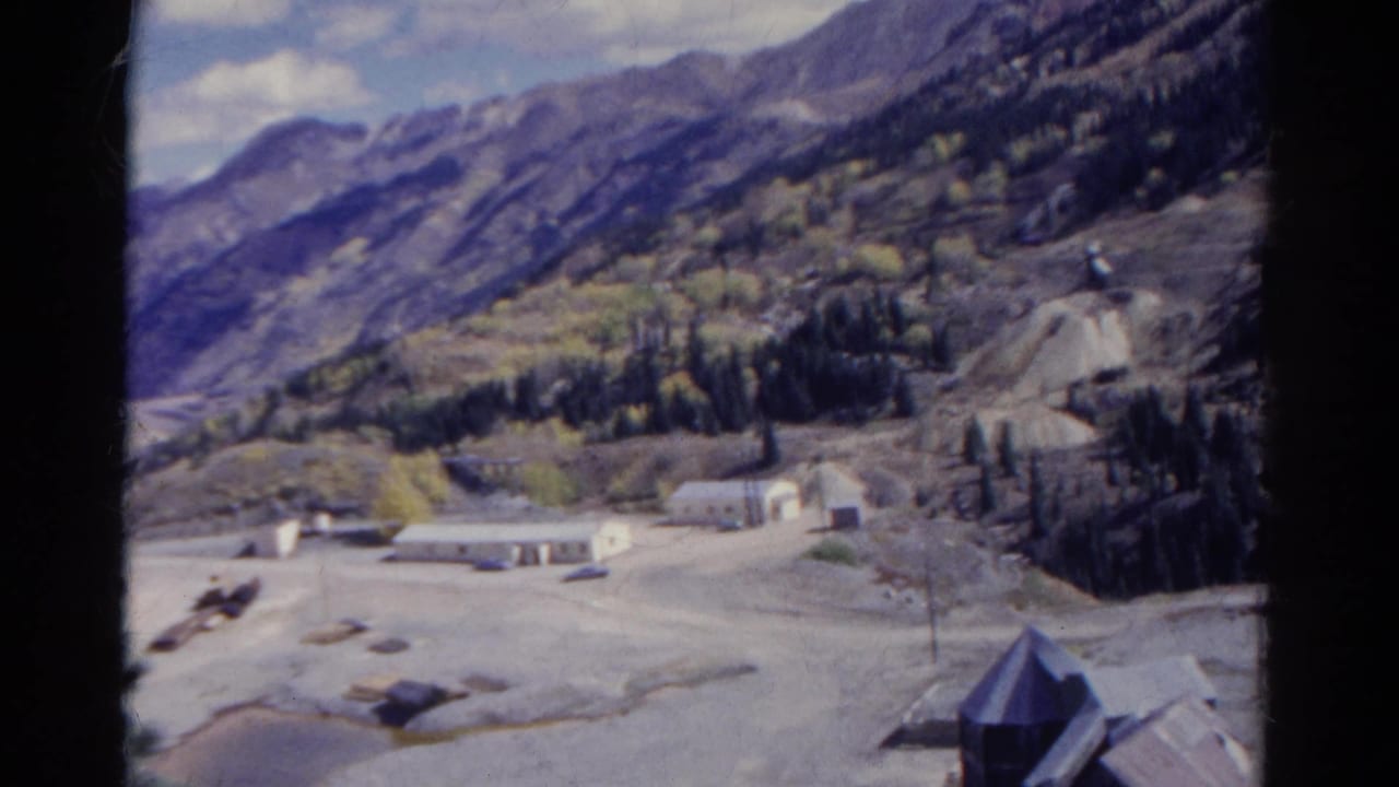 1967 Colorado 8mm frame showing a high view over a mountain valley with scattered buildings, evergreen trees, and pale open ground below rugged slopes.