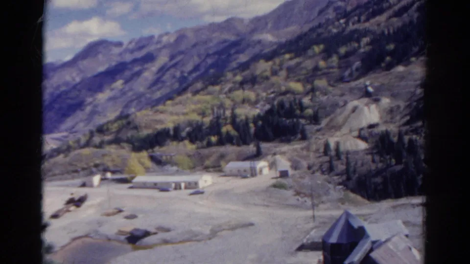 1967 Colorado 8mm frame showing a high view over a mountain valley with scattered buildings, evergreen trees, and pale open ground below rugged slopes.