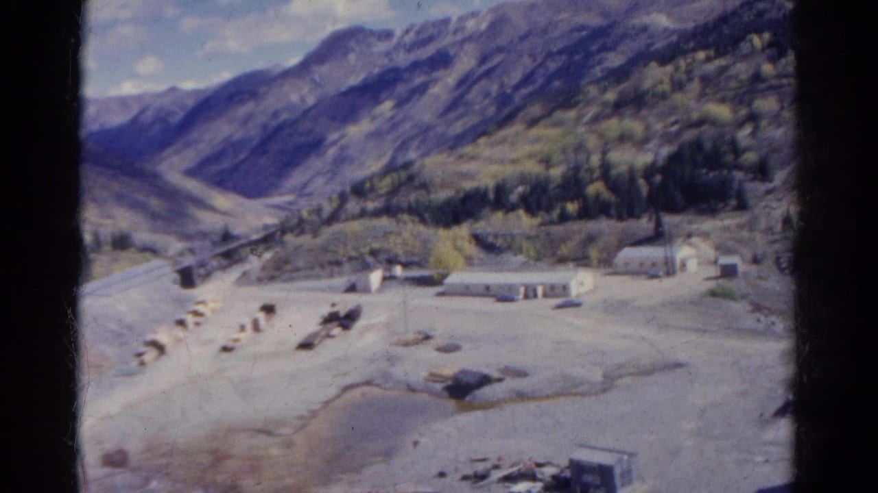 Mountain valley with a winding road, small buildings, trees, and open pale ground.