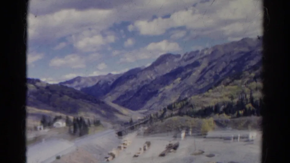1967 Colorado 8mm frame with a broad valley view, low roadside buildings, tree clusters, and mountain ridges under a partly cloudy sky.