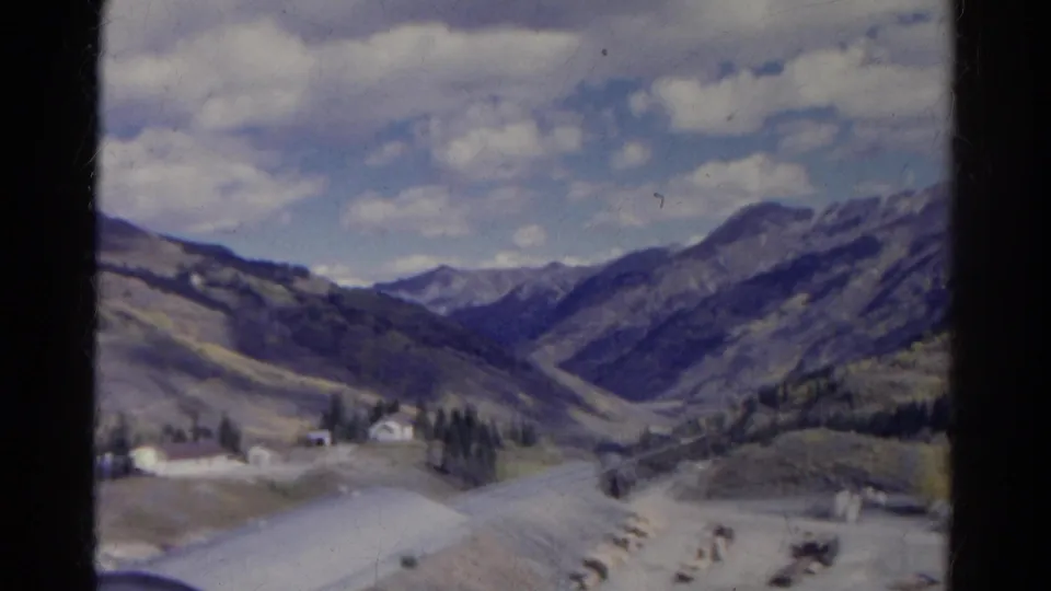 1967 Colorado home-movie frame showing distant peaks, a valley road, scattered buildings, evergreen trees, and pale open terrain.